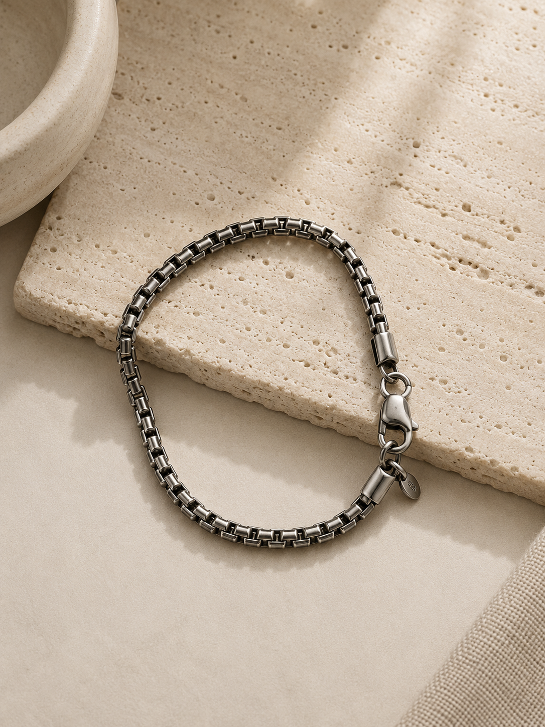 A silver-toned metal chain bracelet with a lobster clasp, styled on a textured beige stone tile and neutral fabric background.