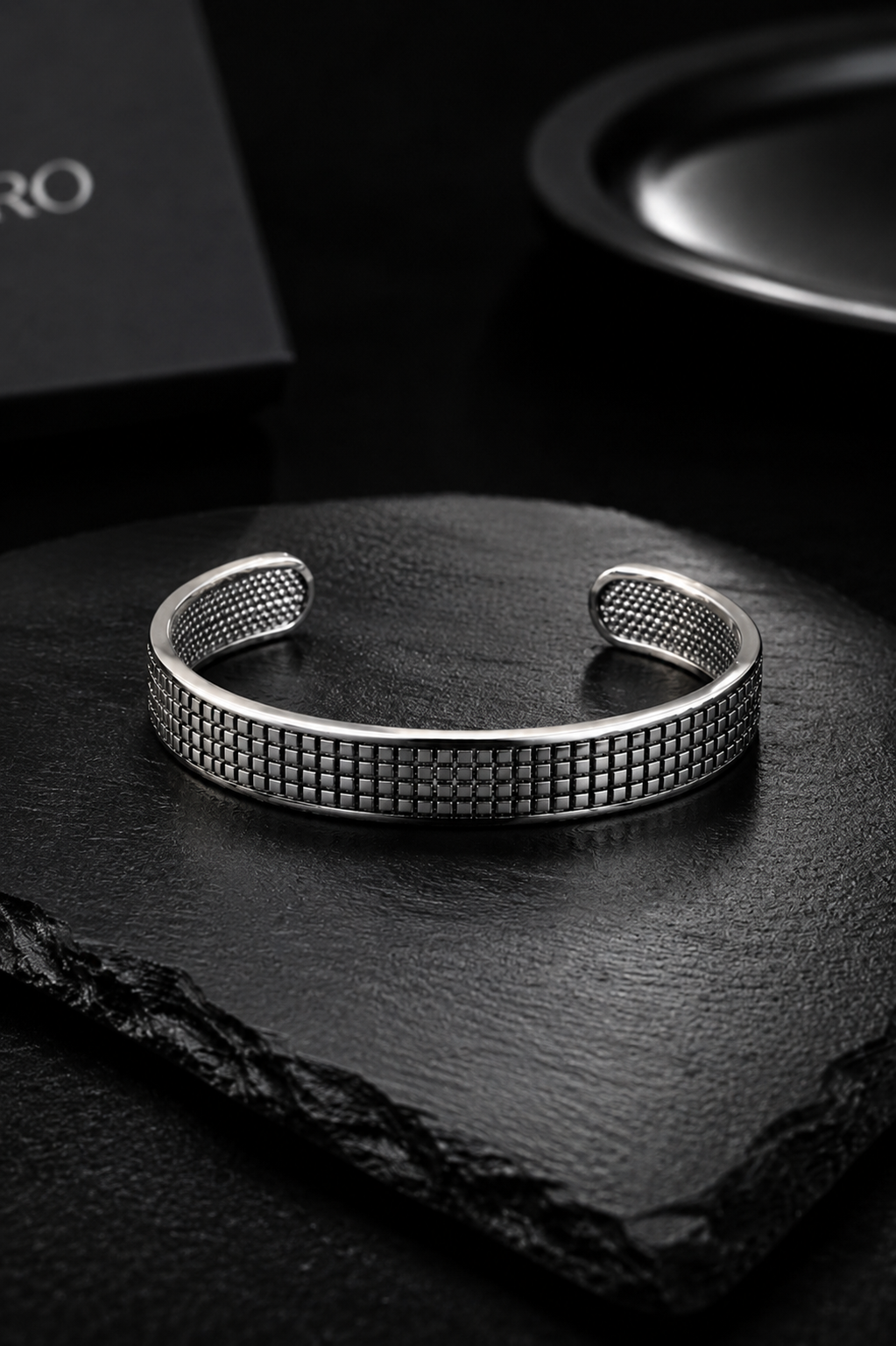 A silver-toned metal cuff bracelet with a textured grid pattern, displayed on a dark slate stone.