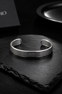 A silver-toned metal cuff bracelet with a textured grid pattern, displayed on a dark slate stone.