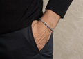A close-up of a man's wrist wearing a silver-toned chain bracelet, dressed in a black ribbed sweater and dark trousers against a neutral beige textured background.