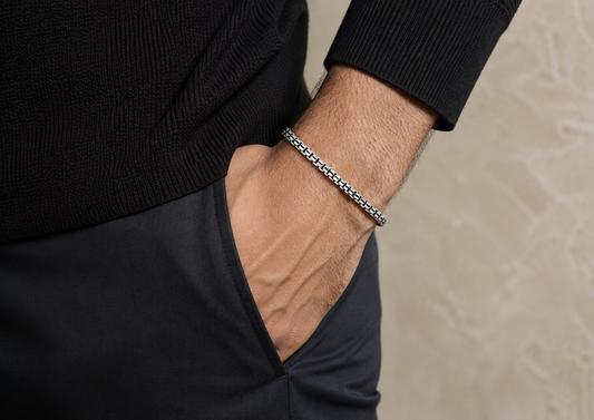 A close-up of a man's wrist wearing a silver-toned chain bracelet, dressed in a black ribbed sweater and dark trousers against a neutral beige textured background.