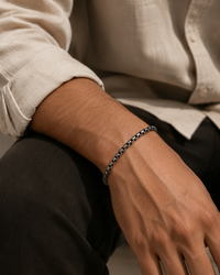 A close-up of a man's wrist wearing a dark metallic chain bracelet, wearing a light beige linen shirt and dark trousers.