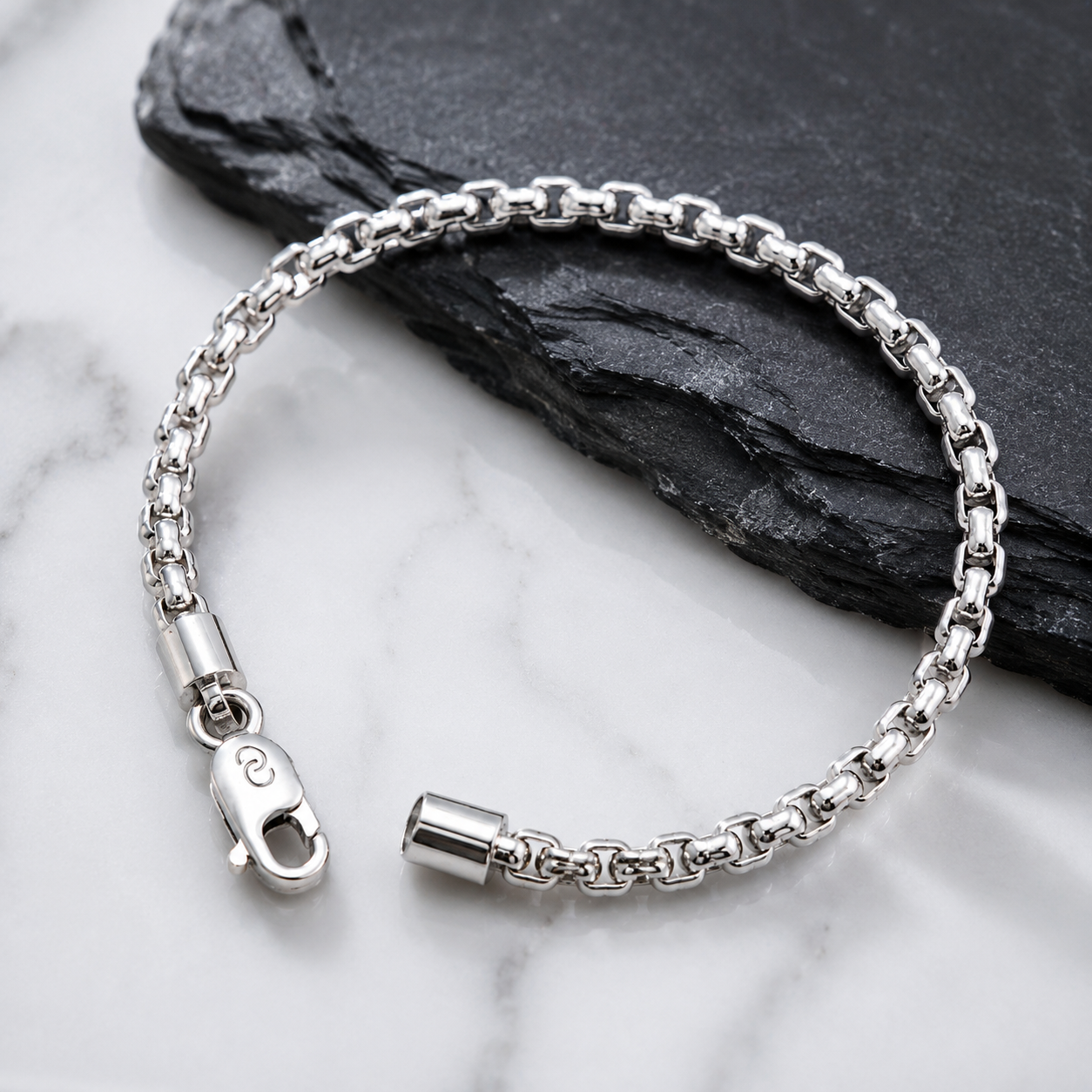 A silver-toned metal chain bracelet with a lobster clasp and cylindrical end cap, arranged on a white marble surface with a dark slate prop.