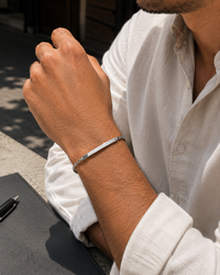 A close-up of a man's wrist wearing a slim silver bar bracelet, dressed in a white linen shirt.