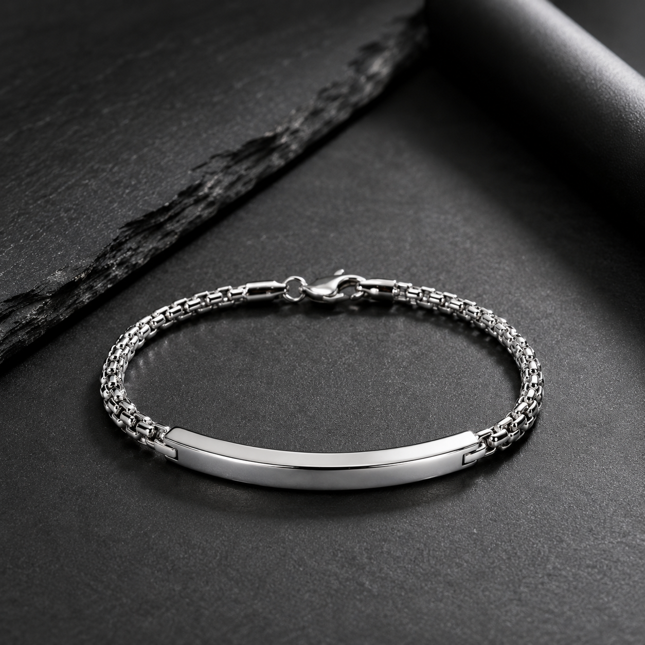 A silver-toned metal bracelet with a smooth central bar and textured chain links, photographed on a dark gray surface with a black stone slab prop.
