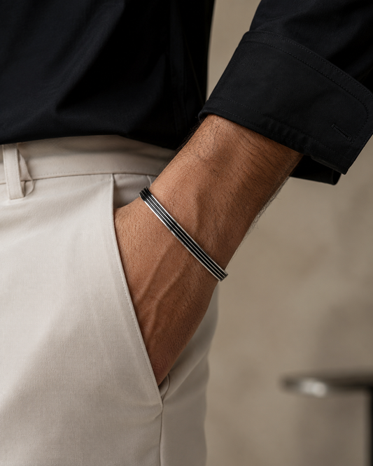 A close-up of a man's forearm wearing a slim three-band silver bracelet, styled with a black shirt and beige trousers.