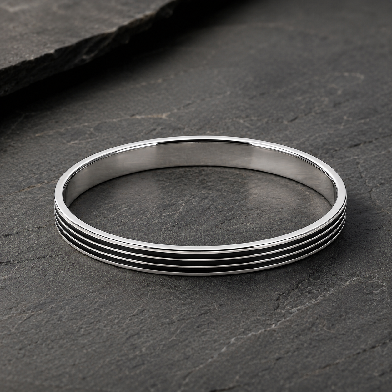 A polished silver-toned bangle bracelet with five parallel grooved lines, displayed on a dark textured slate surface.